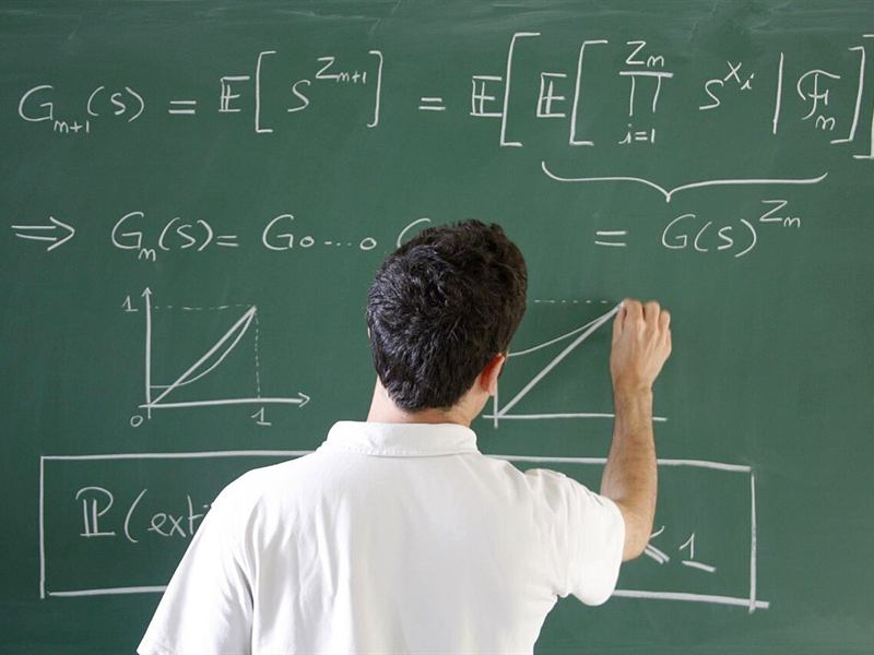 Teacher seen from behind writing equations on a green chalkboard